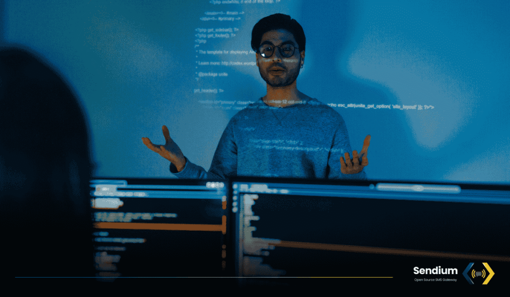 A man wearing glasses stands in a dark room illuminated by blue light from a projector displaying code behind him, with the Sendium Open Source SMS Gateway logo in the bottom right corner.